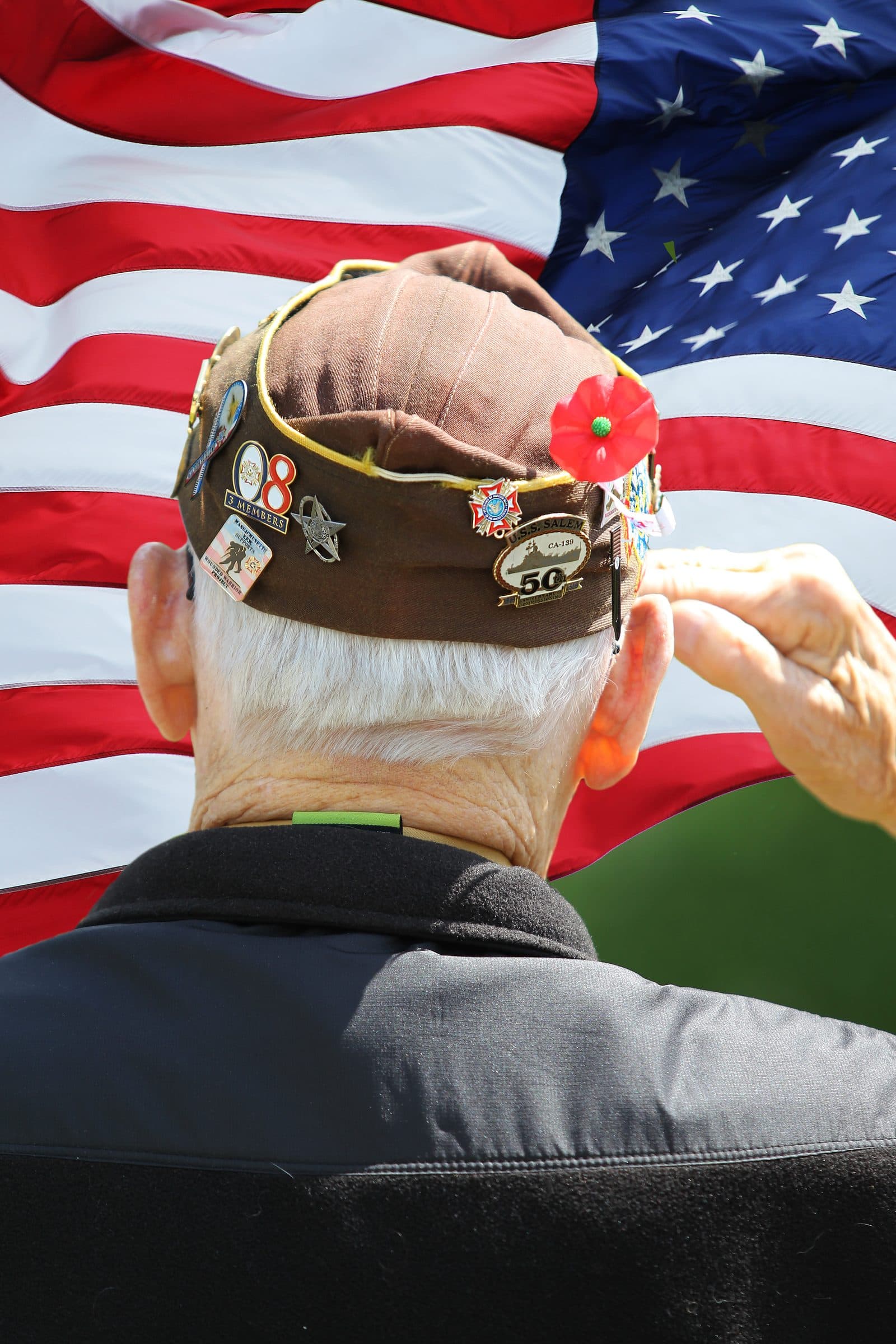 An older veteran in a service cap salutes in front of an American flag